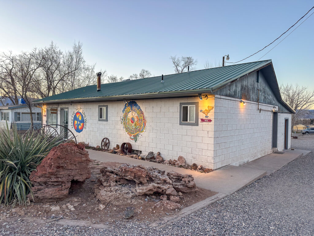 A office building/bath house made of white cinder block with an aluminum roof. There are three windows and a door in the front, with two abstract murals on the front and a sign to the mens bathroom.