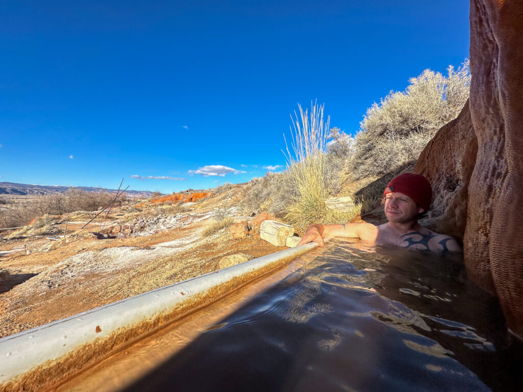 A cast iron soaking tub filled with hot spring water, with a man soaking in the tub in a beanie.