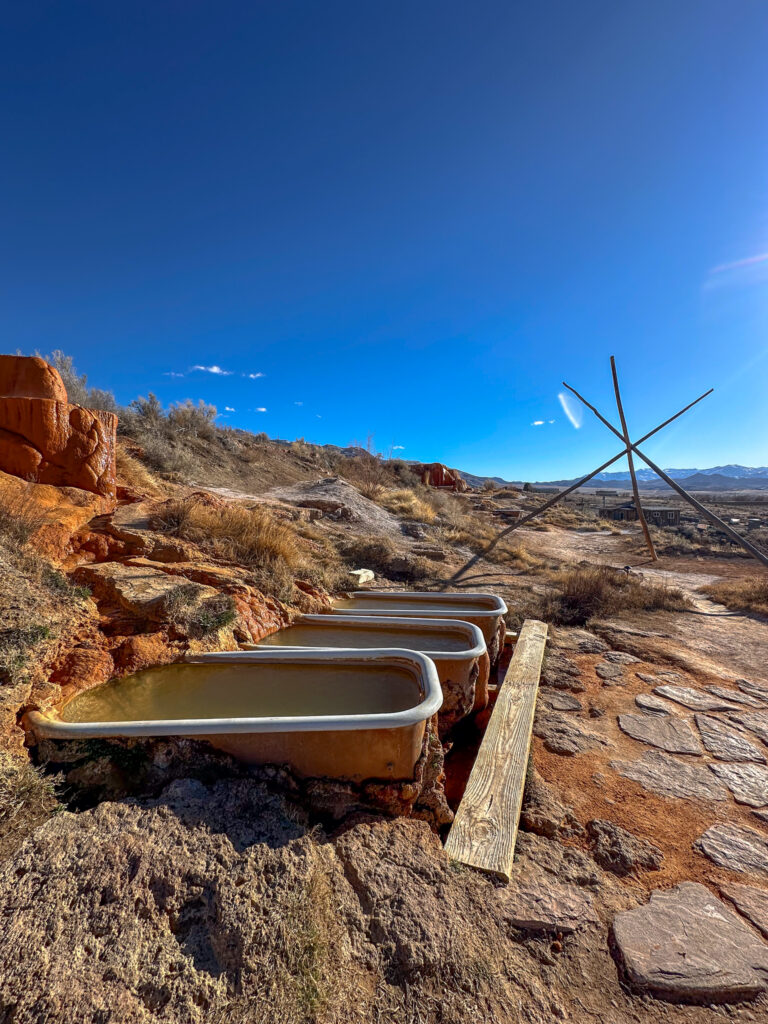 A row of three cast iron tubs filled with hot springs water at Mystic Hot Springs with a blue sky and a desert landscape behind.